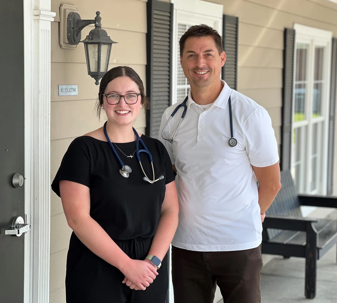 Hannah and Robert Two healthcare professionals, one in a black outfit with a stethoscope and the other in a white polo, stand outside a building.
